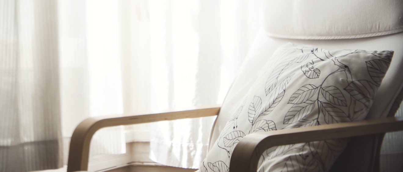 A cozy armchair with a white cushion and a leaf-patterned pillow, placed beside a sheer curtained window with soft light filtering through.