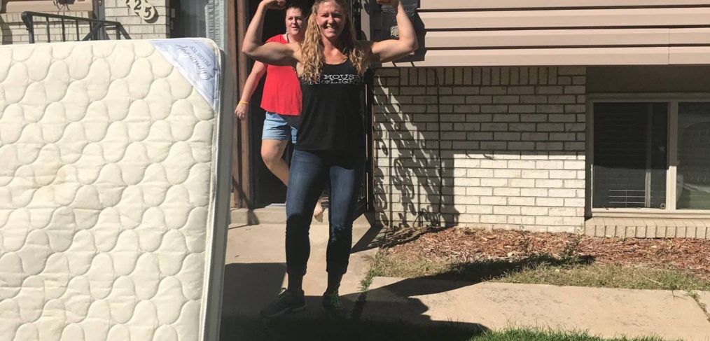 Woman flexing her muscles in front of a house, with a man standing behind her and a mattress leaning on the left.