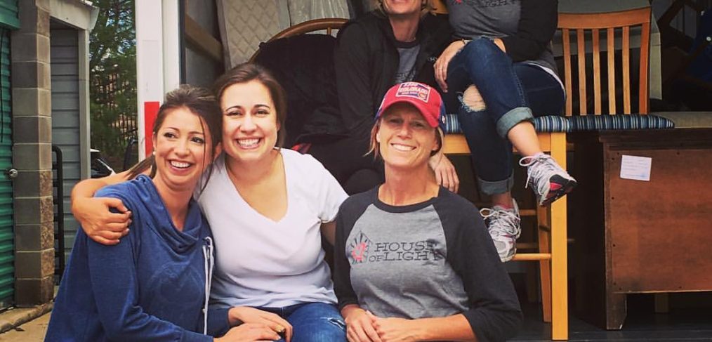 Five women smiling at the camera, sitting and standing in the back of an open moving truck loaded with furniture.