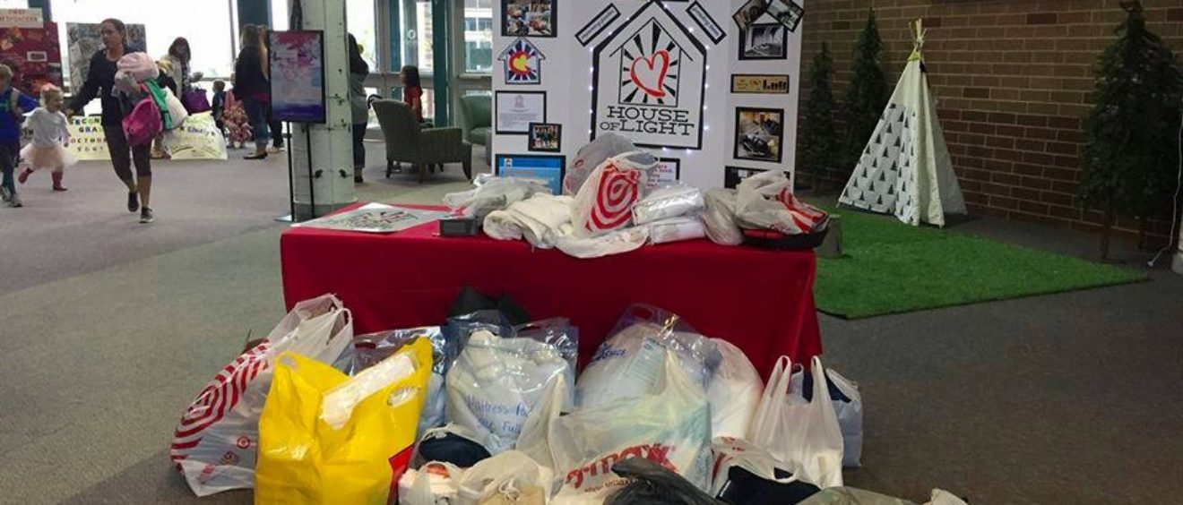 A donation booth with various bags of clothes, a sign reading "house of refuge," and people walking in the background in an indoor setting.