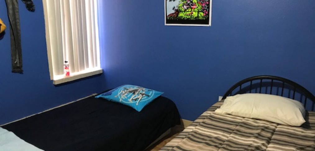 A small bedroom with a single bed covered in striped bedding, a blue wall, a vibrant poster on the wall, and a window with blinds on the left.