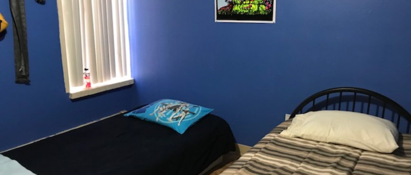 A small bedroom with a single bed covered in striped bedding, a blue wall, a vibrant poster on the wall, and a window with blinds on the left.