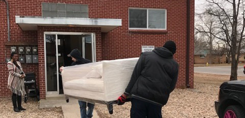 Two people carrying a mattress into a brick building, with a woman holding the door open for them.