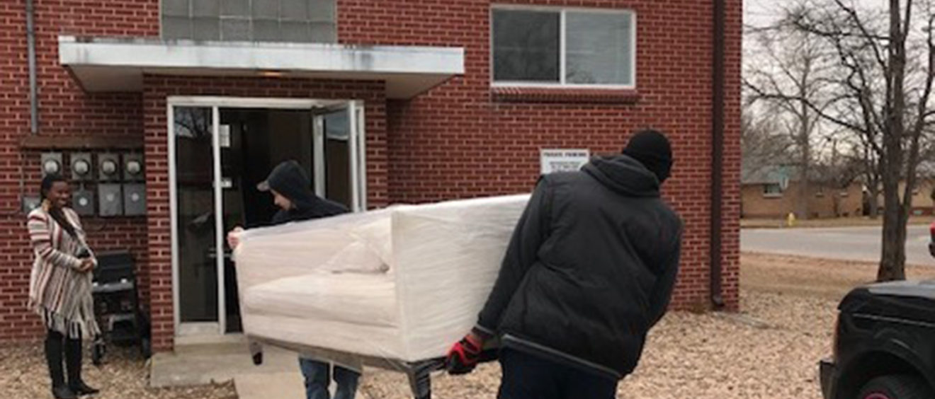 Two people carrying a mattress into a brick building, with a woman holding the door open for them.