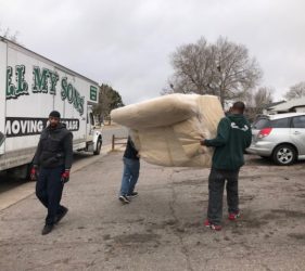 Two movers carrying a large, wrapped piece of furniture towards a moving truck parked on a residential street.