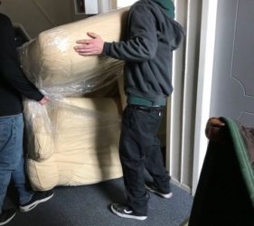 Two men carrying a large, wrapped sofa through a narrow doorway inside a building.