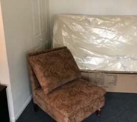 A patterned armchair placed in a corner next to a door and a mattress wrapped in plastic.