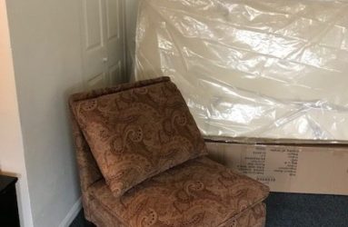 A patterned armchair placed in a corner next to a door and a mattress wrapped in plastic.