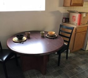 A round dining table with three plates and cups, near a kitchen counter with appliances and a sink, under a bright window.