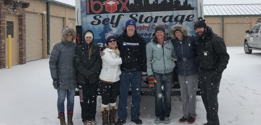 A group of seven adults smiling in front of the "iboxx self storage" sign on a snowy day.