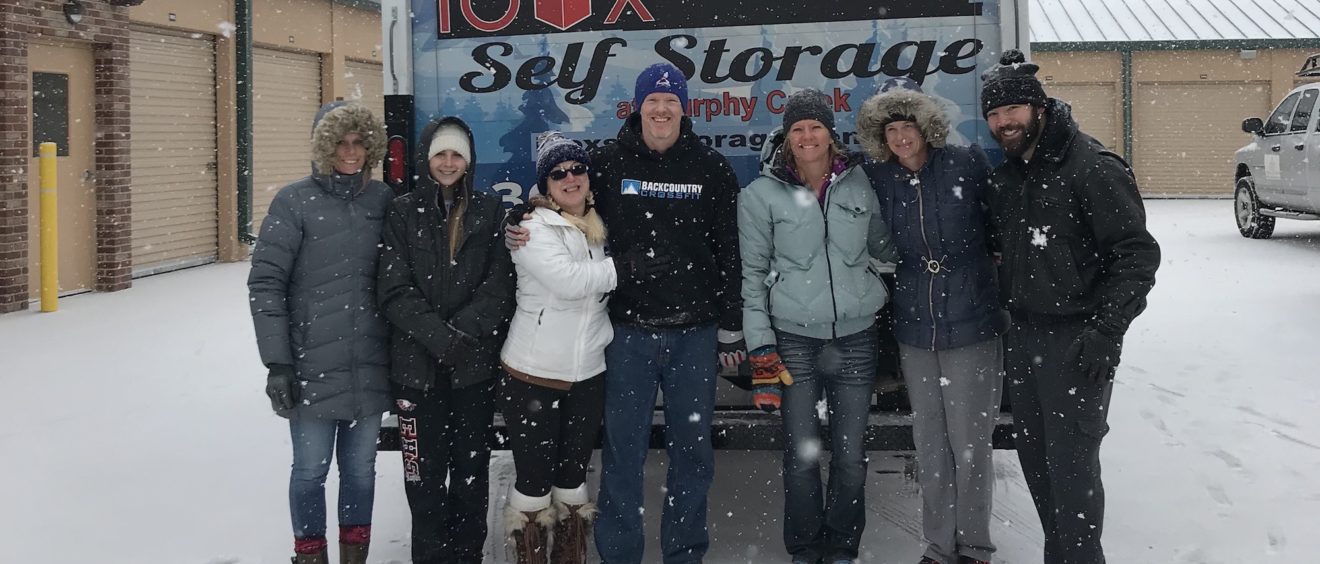 A group of seven adults smiling in front of the "iboxx self storage" sign on a snowy day.