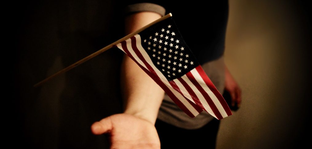 An outstretched hand holding a small american flag on a wooden stick, with a dark, blurred background.
