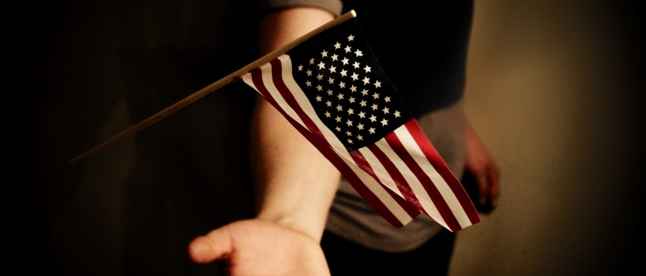 An outstretched hand holding a small american flag on a wooden stick, with a dark, blurred background.
