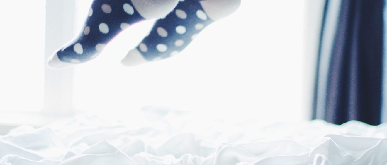 A person wearing polka dot socks with their feet raised above a fluffy white bedspread, backlit by natural light from a window.