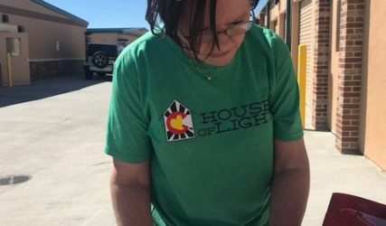 A woman in a green t-shirt labeled "house of light" is writing on papers atop a red clipboard, standing outdoors.