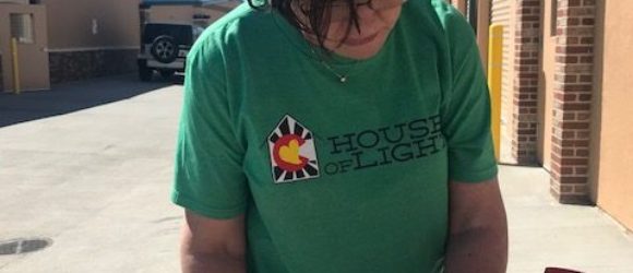 A woman in a green t-shirt labeled "house of light" is writing on papers atop a red clipboard, standing outdoors.