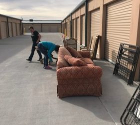 Two people moving a patterned sofa outside a storage unit with various items and open doors in the background.