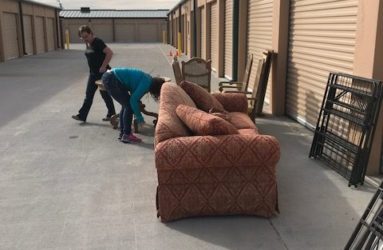Two people moving a patterned sofa outside a storage unit with various items and open doors in the background.