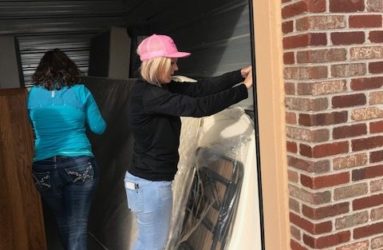 Two women hanging plastic sheeting on a garage door, one wearing a pink hat and the other in turquoise, at a brick building.