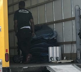 A man wearing a black t-shirt labeled "dew got crew" loads heavy rolled-up mats into the back of a yellow delivery truck.