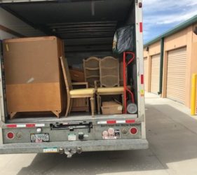A moving truck with its rear door open, revealing furniture and boxes packed inside, parked at a storage facility.