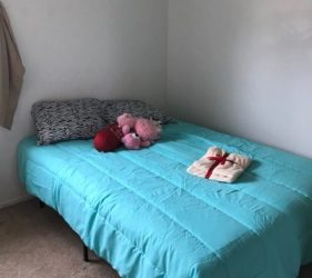 A neatly made single bed with a turquoise bedspread, scattered with a few plush toys and a small wrapped gift, in a plain, light-colored room.