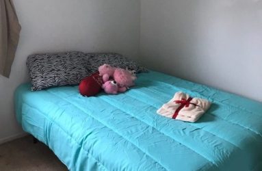 A neatly made single bed with a turquoise bedspread, scattered with a few plush toys and a small wrapped gift, in a plain, light-colored room.