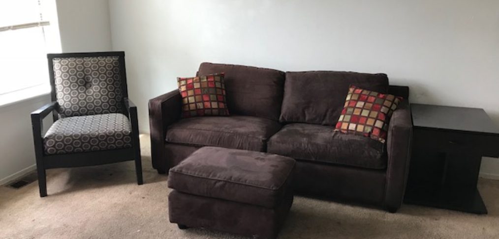 A living room with a brown couch, matching ottoman, a patterned armchair, and a black side table near a window.