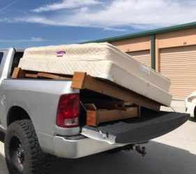 A white pickup truck with its tailgate open, loaded with a disassembled wooden bed frame and a folded mattress in a storage facility parking lot.