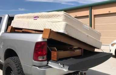A white pickup truck with its tailgate open, loaded with a disassembled wooden bed frame and a folded mattress in a storage facility parking lot.
