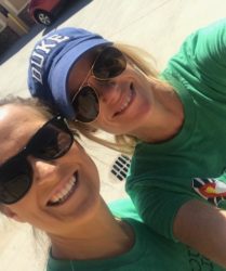 Two women in green t-shirts and sunglasses smiling for a selfie, one wearing a baseball cap with "duke" on it, in a sunny parking lot.