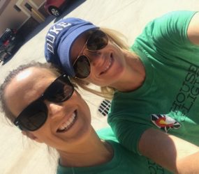 Two women in green t-shirts and sunglasses smiling for a selfie, one wearing a baseball cap with "duke" on it, in a sunny parking lot.