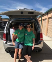 Two women in green t-shirts standing in front of an open car trunk, smiling, in a sunny storage facility area.