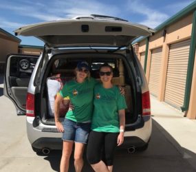 Two women in green t-shirts standing in front of an open car trunk, smiling, in a sunny storage facility area.