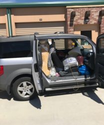 A silver minivan with open doors, packed with various items including bags, clothing, and containers, parked in front of a storage unit.