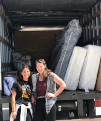 Two women standing in front of an open moving truck loaded with furniture and mattresses, smiling at the camera.