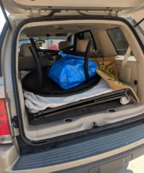 Car trunk open, containing a black suitcase, a blue trash bag, a plank, and a white foam sheet, parked outdoors in daylight.