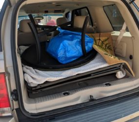 Car trunk open, containing a black suitcase, a blue trash bag, a plank, and a white foam sheet, parked outdoors in daylight.