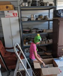 A person in a frog hat sits by a box in a cluttered garage, surrounded by shelves filled with various items.