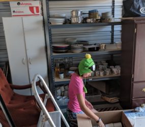 A person in a frog hat sits by a box in a cluttered garage, surrounded by shelves filled with various items.