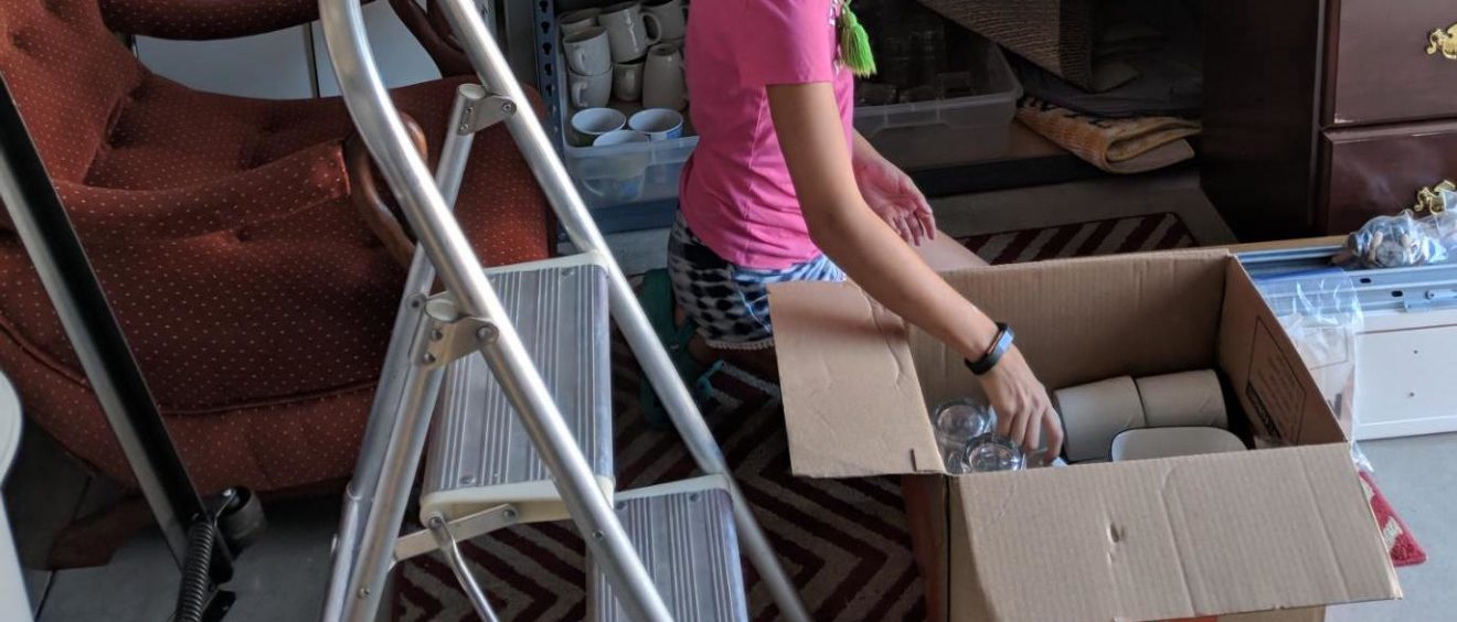 A woman in a pink shirt uses a walker while packing glassware into a cardboard box in a cluttered room.