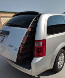 Rear view of a white dodge grand caravan with an open trunk, filled with a plaid suitcase, parked under sunlight, showing some damage on the bumper.