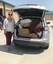 A woman standing beside a car with its trunk open, filled with packed boxes and bags, outside a storage facility.