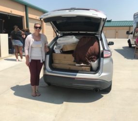 A woman standing beside a car with its trunk open, filled with packed boxes and bags, outside a storage facility.