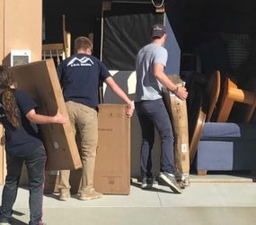 Three people loading cardboard boxes and furniture into a truck outside a building.
