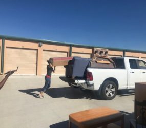 Two people loading furniture into a pickup truck in front of storage units on a sunny day.
