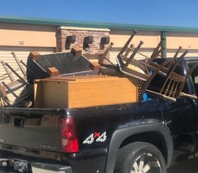 A black chevrolet pickup truck loaded with assorted furniture including chairs and wooden pieces, parked outside storage units.