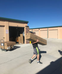 A person carrying a large cardboard box outside a storage facility on a sunny day.