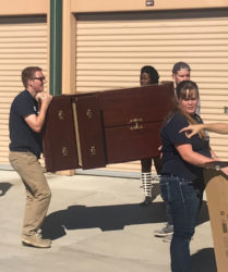 Group of people moving furniture outdoors, one man carrying a wooden cabinet while others assist and coordinate.