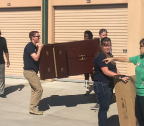 Group of people moving furniture outdoors, one man carrying a wooden cabinet while others assist and coordinate.